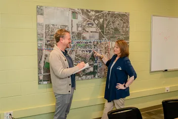 A staff member points at a large wall-mounted campus map while another takes notes. They appear to be discussing safety logistics.