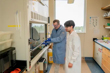 Two lab workers in protective coats and goggles stand at a fume hood as one demonstrates proper usage and safety protocol.