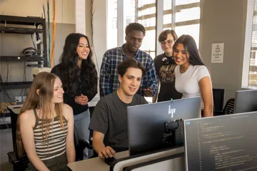 A group of students looking at a students desktop while they work on it