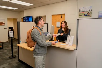Inside the Bursar’s Office at CSUMB, a student makes a cash payment to a smiling finance staff member across the front counter.