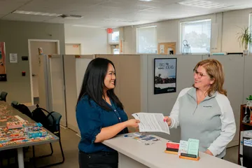 A ���ӽ紫ý HR representative smiles while handing an employee paperwork across a front desk in a bright and welcoming office, decorated with plants and inspiration posters.