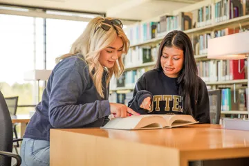 Two students sit together at a library table, smiling and pointing at a book as they study and collaborate.