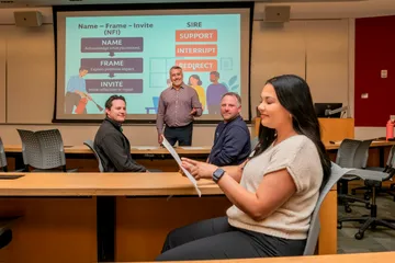 CSUMB Ombuds standing at the front of a classroom while faculty members review printed materials, with a presentation slide about communication strategies projected behind them.
