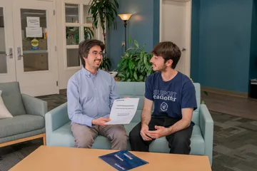 An HR employee and a CSUMB student sit on a couch in the HR reception area reviewing a new hire packet for student assistants, surrounded by welcoming decor and indoor plants.