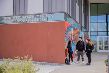 A group of three people is talking together. They are standing in front of a painted wall, with a CAHSS building nearby.