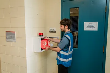 The same student retrieves an AED (defibrillator) from a wall-mounted emergency case outside the Safety, Risk & Sustainability office door.