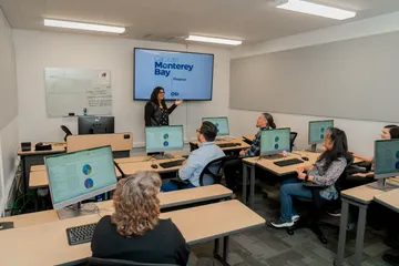 A CSUMB finance staff member presents to colleagues in a training room while standing in front of a display reading 