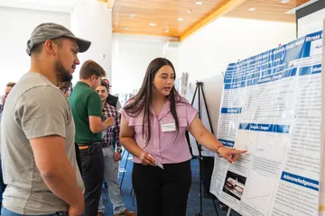 A student at CSUMB explains a research poster on agricultural science to a group of attendees during the AGPS Summer Presentation. The student gestures toward the poster while others listen closely.