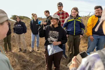 A group of students stands in a circle with a women in the middle holding up a field mouse.