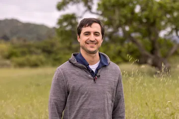 Portrait of a smiling AES faculty member amidst a backdrop of grass and trees.