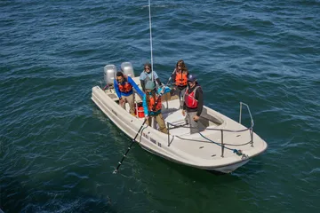 A group of four students and a professor on a boat with life jackets on lowering a pole into the ocean