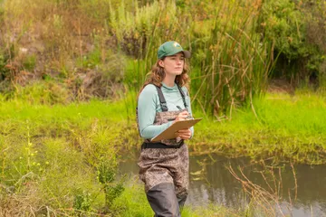 An environmental science student standing in grass next to a body of water writing on a clipboard
