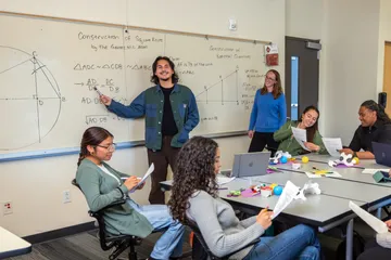 Student standing near white board teaching other students who are sitting at a table