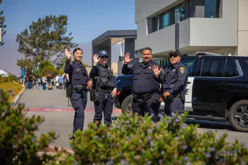 Four CSUMB UPD officers stand in front of a police SUV, waving and smiling at the camera. The Otter Student Union and passing students appear in the background.