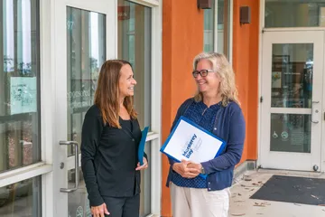 Two staff members stand outside the Sponsored Programs Office entrance, chatting and smiling while holding CSUMB-branded folders.