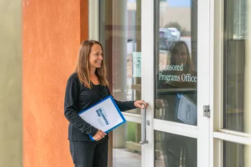 A staff member opens the door to the Sponsored Programs Office while holding a CSUMB Sponsored Programs folder and smiling.