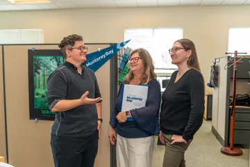 Three team members stand inside the Sponsored Programs office near decorated cubicles, chatting casually under a CSUMB pennant.