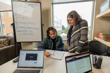 A student explaining a question to a professor who is standing next to him helping him on a paper.