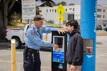 A ���ӽ紫ý parking officer smiles while pointing at a kiosk screen, assisting a student. A Parking Services vehicle and signage are visible in the background.