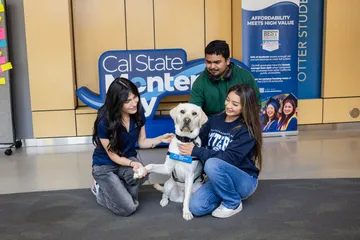 Three students kneel and sit around the CSUMB service dog, smiling and petting it in front of a 