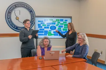 Four ���ӽ紫ý staff members collaborate in a meeting room with a digital display showing a flowchart of university functions. One person points at the screen while others smile and interact around a laptop.