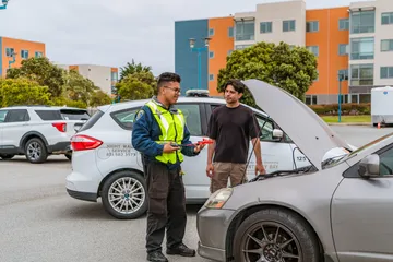 A CSUMB community service officer in a yellow vest explains how to use jumper cables while assisting a student with a stalled car. The Night Walk Service vehicle is behind them.