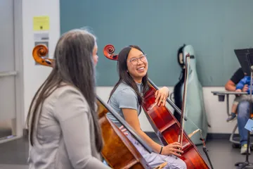 A ���ӽ紫ý cello student laughs warmly during a lesson, sharing a lighthearted moment with her instructor while holding her instrument.