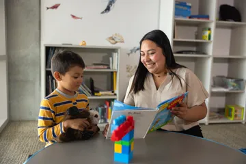 A child, clutching teddy bears, looks at a book a woman is showing him.