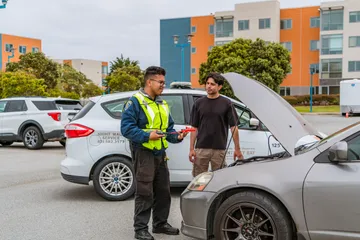 A CSUMB community service officer in a yellow vest explains how to use jumper cables while assisting a student with a stalled car. The Night Walk Service vehicle is behind them.