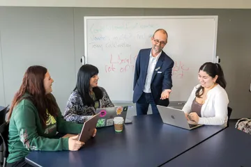Professor stands at a table engaging with three students using laptops in a classroom setting