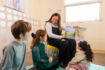 Student reading a book to three children sitting on the ground.