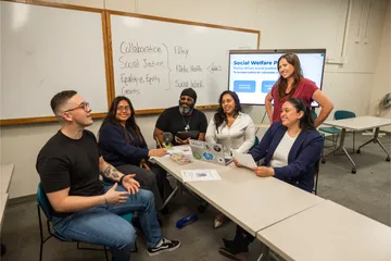 Five students sitting at a table listening while one of them is speaking to the professor who is standing smiling