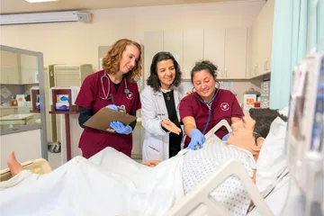 A group of two nursing students in scrubs and a professor using a stethoscope on a training dummy and writing down results