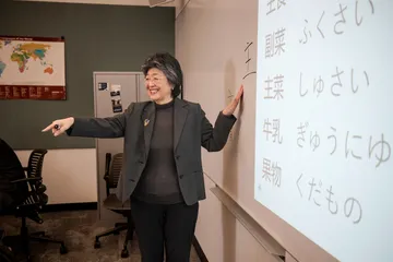 Professor standing in front of a white board pointing at Japanese symbols and smiling while teaching the class