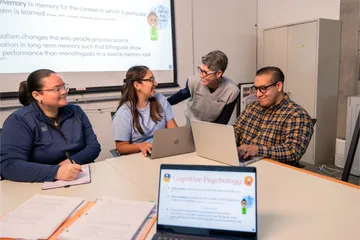 Three professors sitting at a table on their computer and taking notes while discussing with a student also sitting at the table