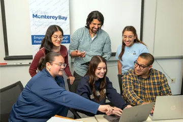 Three students sitting at a table looking at the a computer in the middle of them, with three students standing behind them also looking at the same computer.