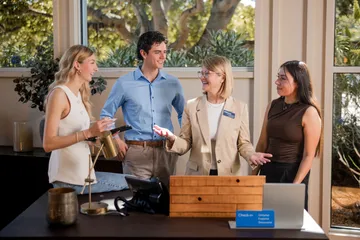Three students standing behind the check in desk with a professor standing in the middle of them with her arms a little extended and smiling