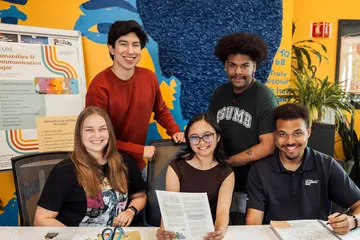 A group of students gather around a table inside a brightly colored campus space, smiling toward the camera while holding papers and notebooks, with humanities and communication program posters and artwork visible behind them.