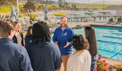 A group gathers by a coastal pool as a staff member leads a conversation in a scenic, community-focused setting