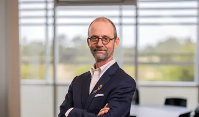 Professor in a suit poses confidently with arms crossed in a bright classroom.