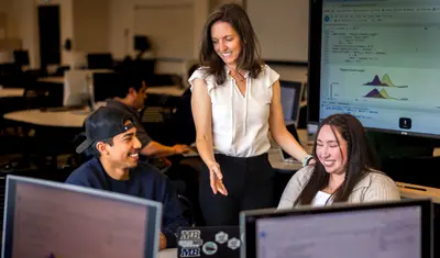 Two students are smiling as they listen to their teacher, who is standing.