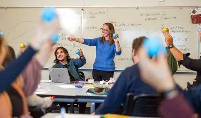 A classroom full of mathematics students tosses plastic balls around in front of a whiteboard