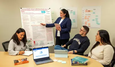 A student presenting a poster of work to three other students who are sitting at a table listening and taking notes.