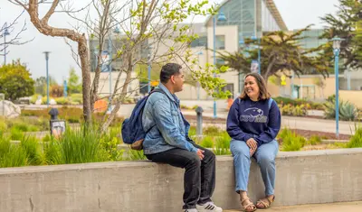 Two people sit outside on campus, smiling and chatting together on a sunny day.