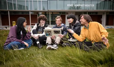 A group of people is sitting on the grass in front of the RND Amphitheatre, looking at a book and smiling.