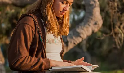 A student reads outdoors beneath a tree, reflecting a peaceful and focused learning moment in a natural setting.