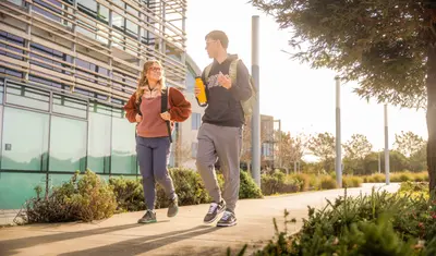 Two CSUMB college students walk and chat on a sunny campus path, carrying backpacks and a water bottle near a modern building.