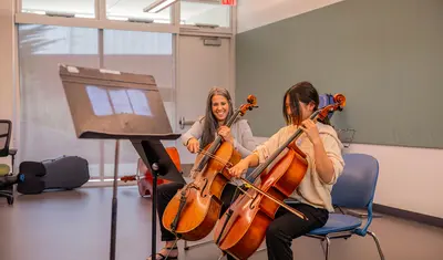 A student and instructor practice cello together, highlighting mentorship and hands-on learning in a music classroom.