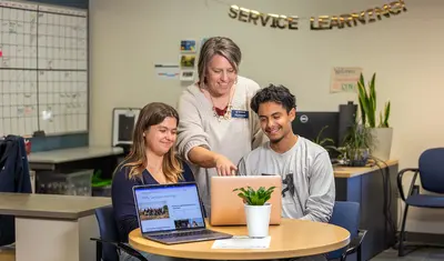Staff member guiding two students through a service-learning project at a table.