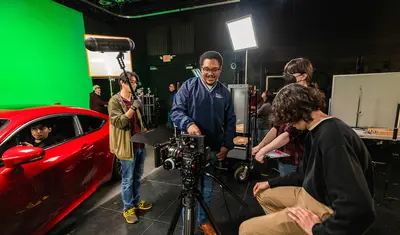 CSUMB Cinematic Arts students work together on a film set in a studio, adjusting a professional camera while others handle audio, lighting, and a red car scene in front of a green screen.